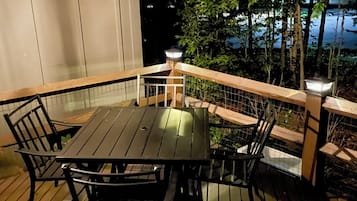 Eine Terrasse im Freien mit Tisch und Stühlen, umgeben von einem Holgitter und Blick auf Bäume sowie ein Gewässer.