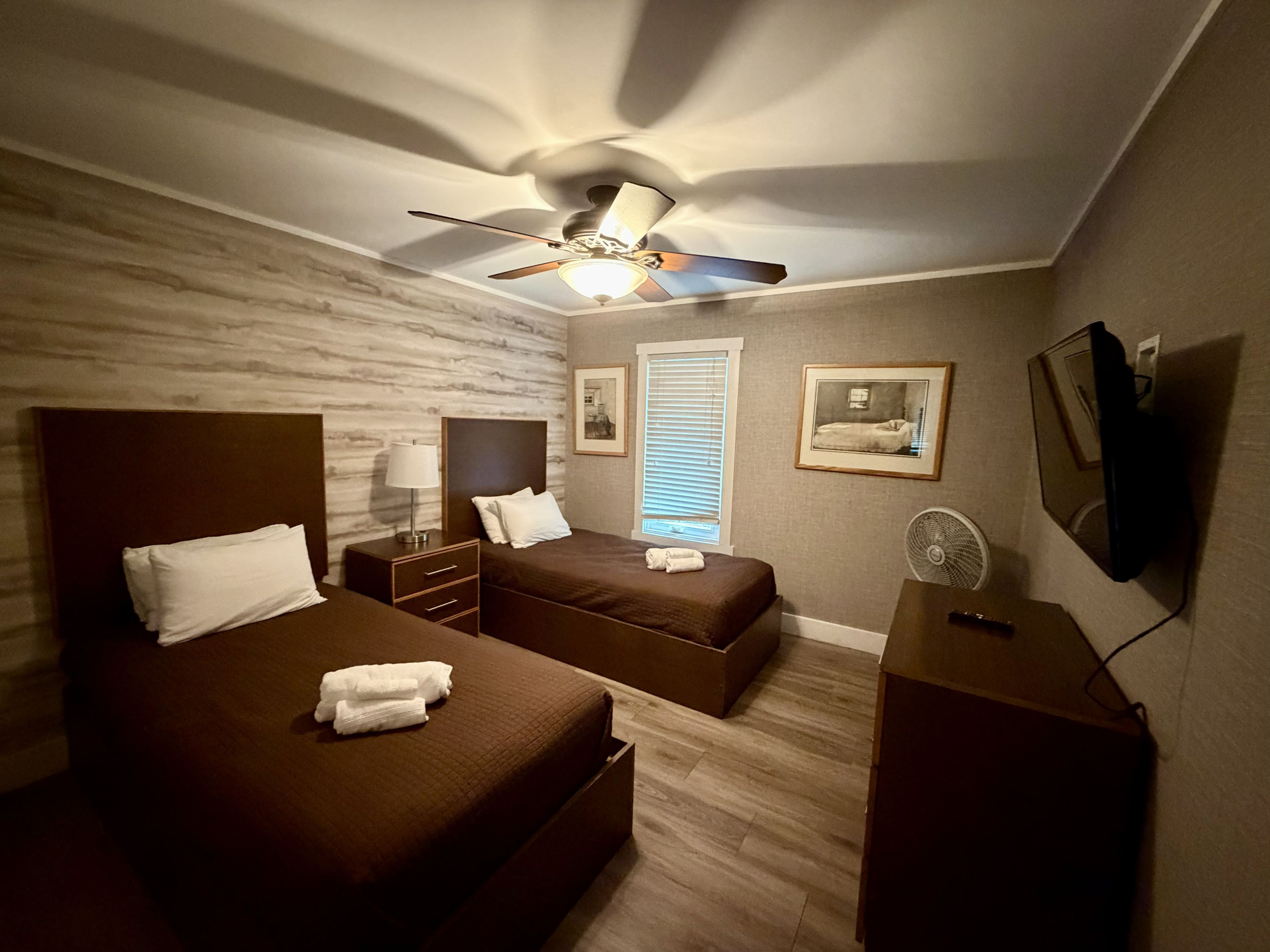 Ein Hotelzimmer mit zwei Betten, einem Deckenventilator, einem Nachttisch, einem Fernseher und einem Fenster mit Jalousien.