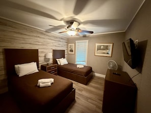 Hotellihuone, jossa on kaksi sänkyä, kattotuuletin, yöpöytä, televisio ja ikkuna, jossa on sälekaihtimet.