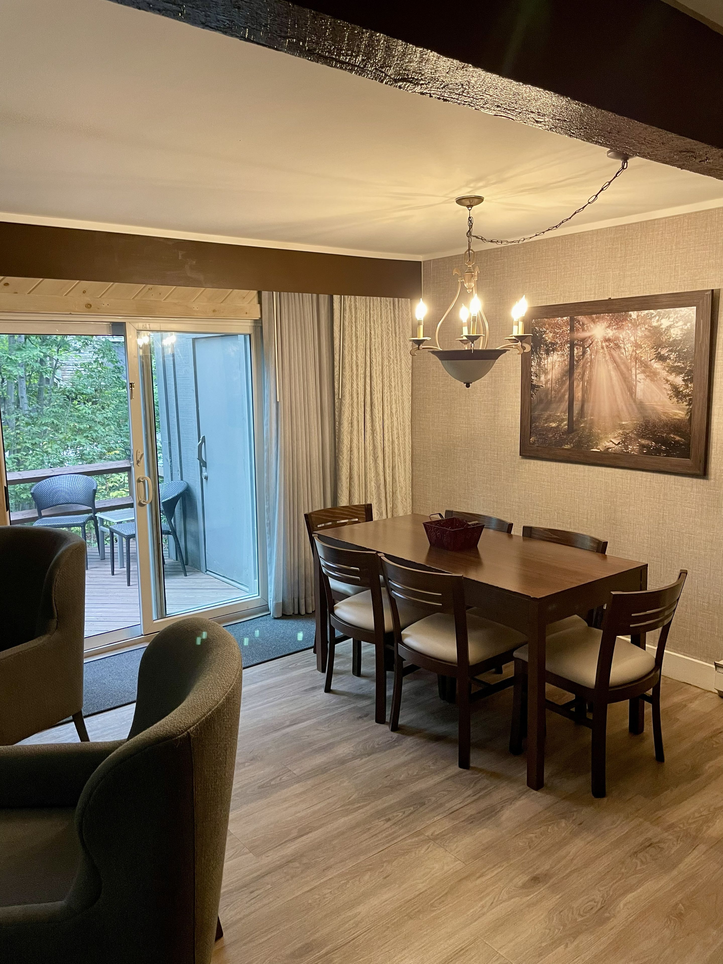 A dining area with a wooden table, chairs, and a chandelier. There is a sliding glass door leading to an outdoor patio with chairs.