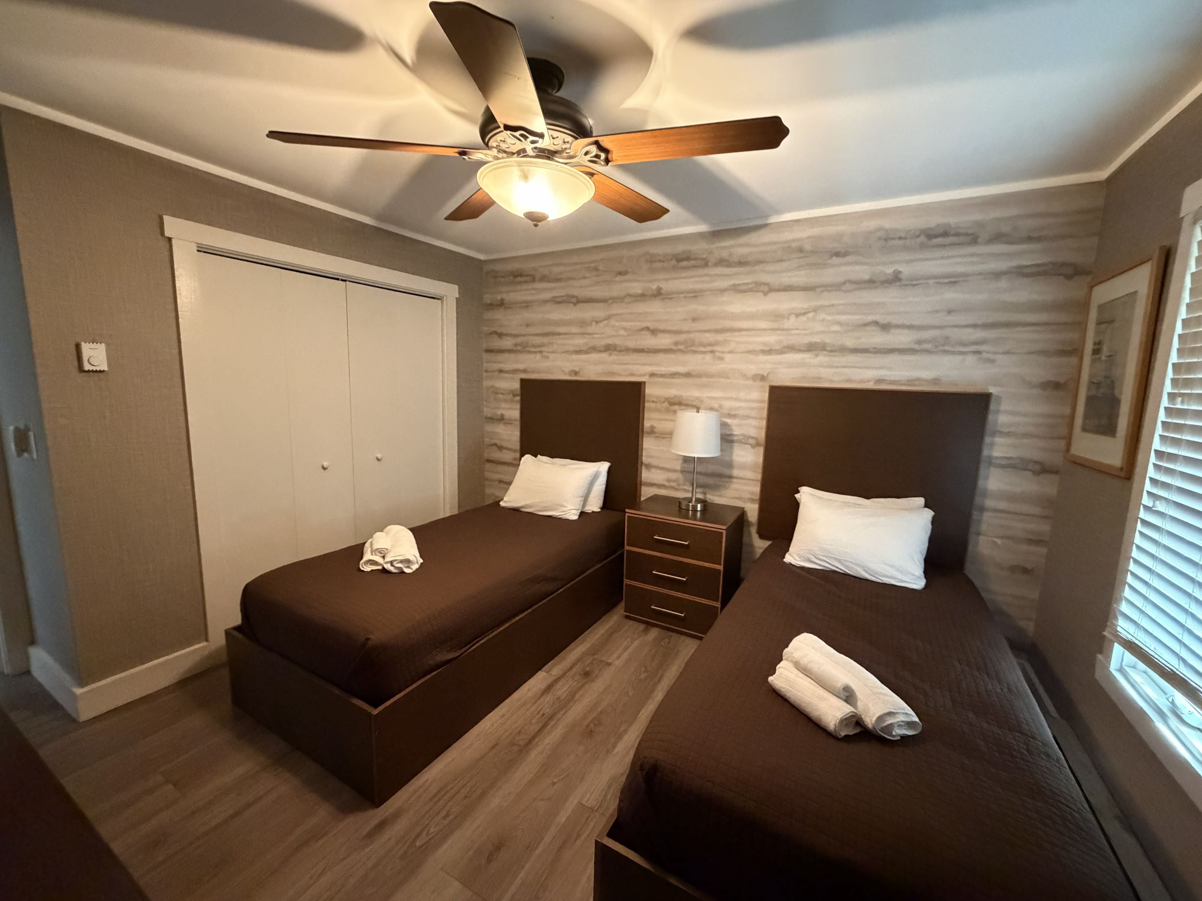 Ein Hotelzimmer mit zwei Betten, einem Deckenventilator, einem Nachttisch mit Lampe und einem Fenster mit Jalousien.