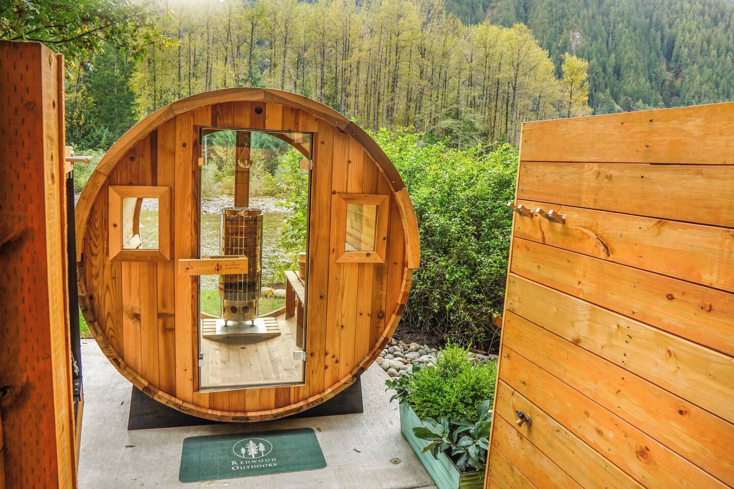 A wooden sauna with a glass door, surrounded by a wooden deck and a forest view.