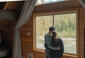 Una pareja abrazándose en una cabaña con un ventanal grande que da a un área boscosa.