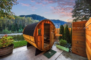 A wooden sauna structure with a curved roof, situated outdoors with a view of a forested hillside and a river in the background. - Special 1-Night Stays The Gateway (Gold Bar)