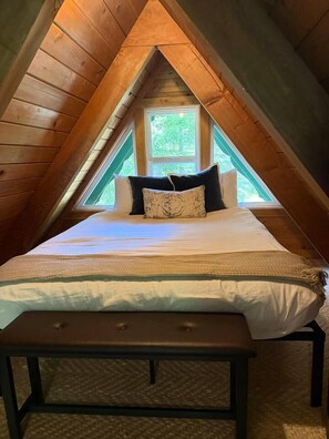 A bedroom with a sloped ceiling, a bed with pillows, a bench, and a window providing a view of greenery outside. - Special 1-Night Stays The Gateway (Gold Bar)
