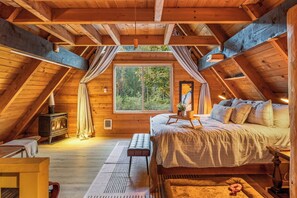 Un dormitorio acogedor en el ático, con techo de madera, una cama, una mesita y una ventana con vistas a follaje.