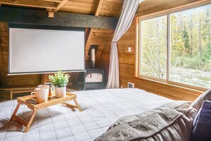 A cozy cabin room with a bed, a projector screen, a fireplace, and a window with a forest view.