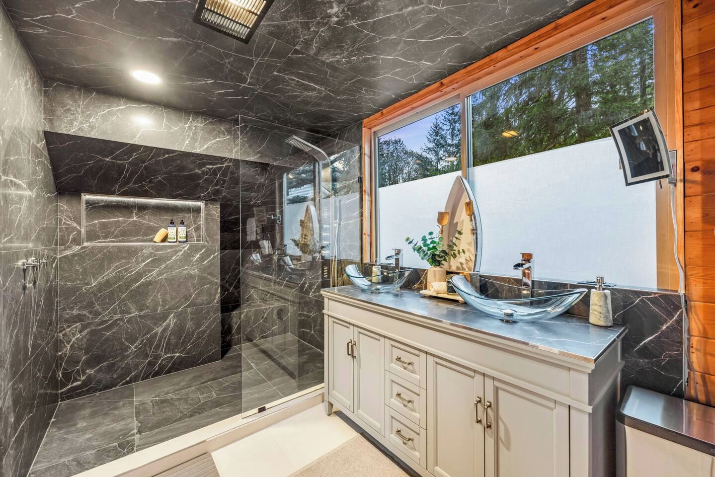 A bathroom with a glass-enclosed shower, marble walls, a wooden-framed window, and a vanity with a large mirror.