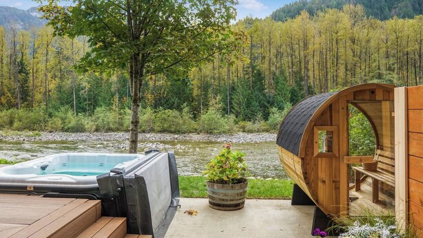 Ein Whirlpool und eine Holzhütte an einem Fluss mit Wald im Hintergrund.