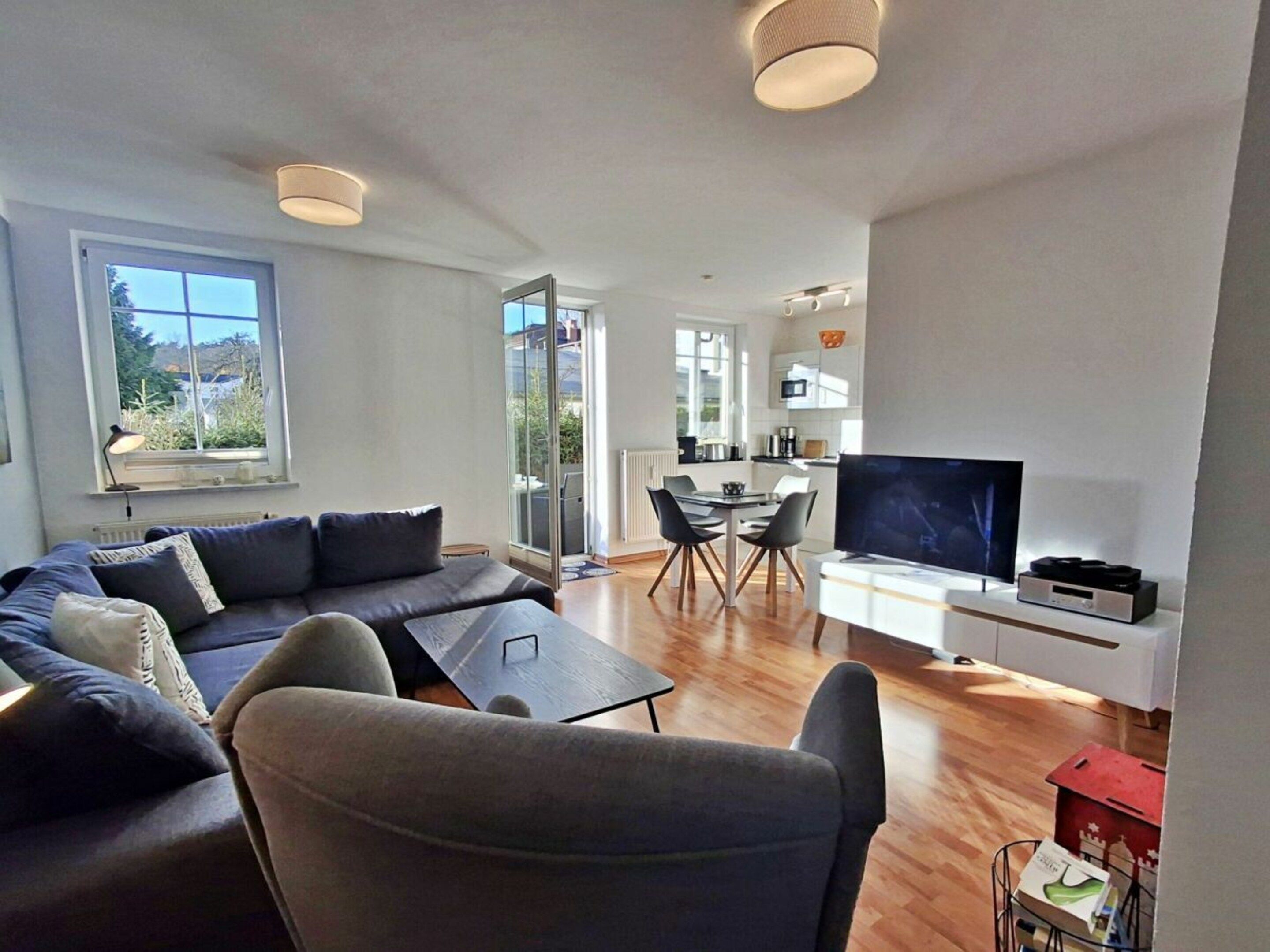 Ein modernes Wohnzimmer mit einer Couch, einem Couchtisch und einem Fernseher. Es gibt einen Essbereich mit Tisch und Stühlen. Das Zimmer verfügt über große Fenster und einen Blick ins Freie.