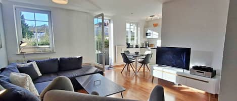 A modern living room with a sofa, a coffee table, and a TV. There is a dining area with a table and chairs. The room has large windows and a view of the outdoors.