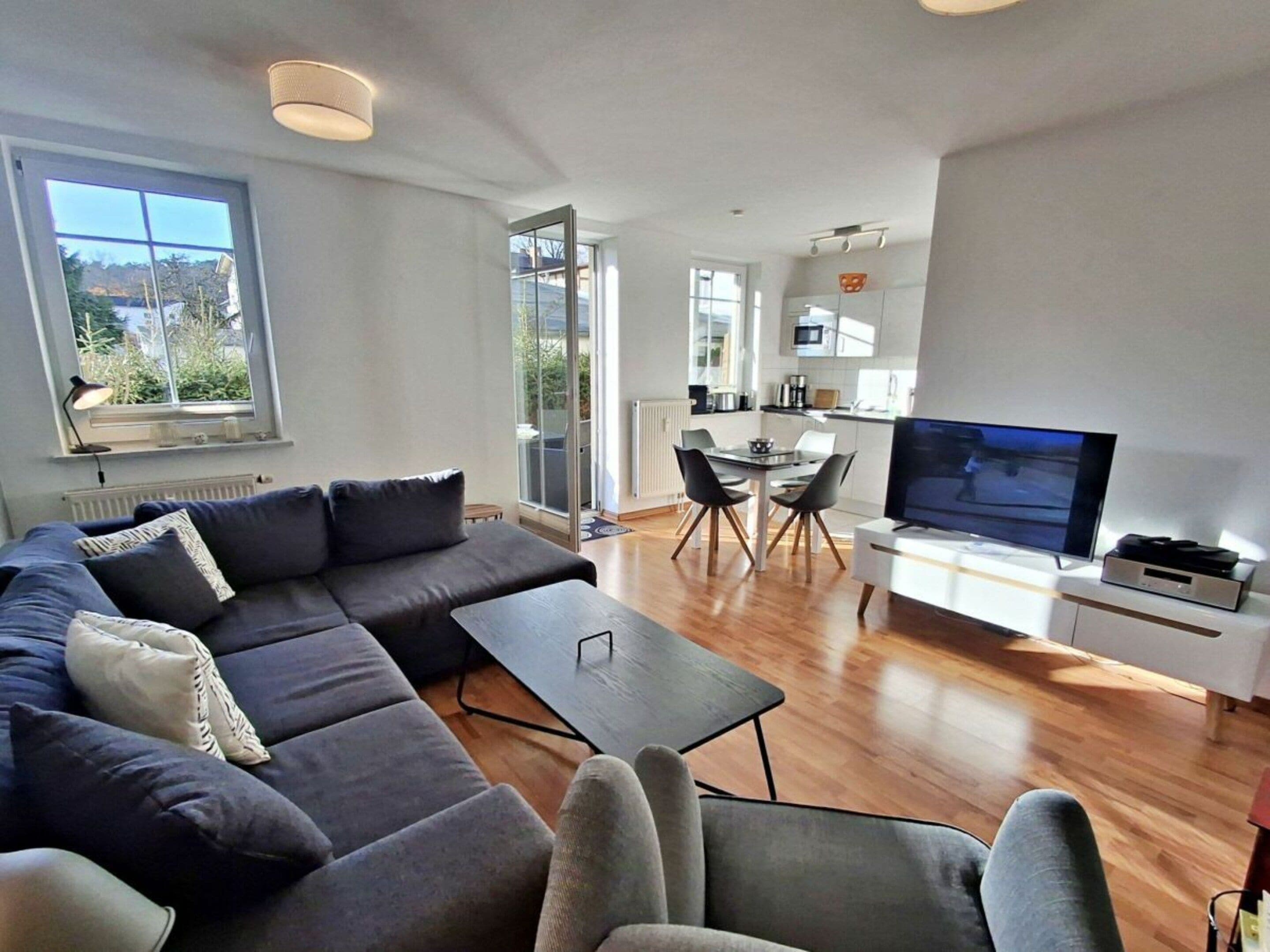 Un salon moderne avec un canapé, une table basse et un téléviseur. Il y a également un coin repas avec une table et des chaises. La pièce est dotée de grandes fenêtres et offre une vue sur l’extérieur.