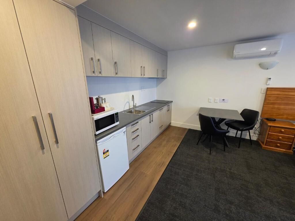 A compact kitchen with white cabinets, a black countertop, and a small refrigerator. There is a dining area with a table and chairs.