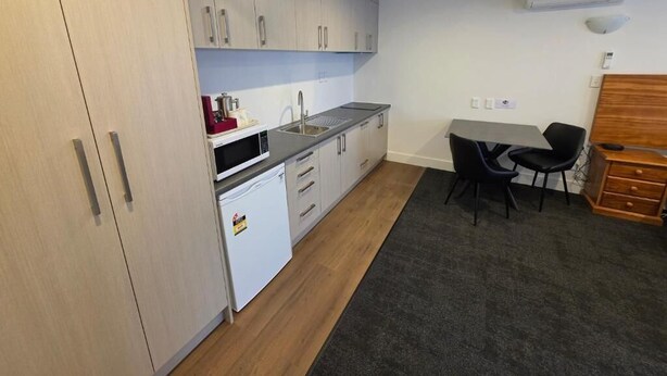 Cocina compacta con armarios blancos, encimera negra y un refrigerador pequeño. Hay una zona de comedor con mesa y sillas.