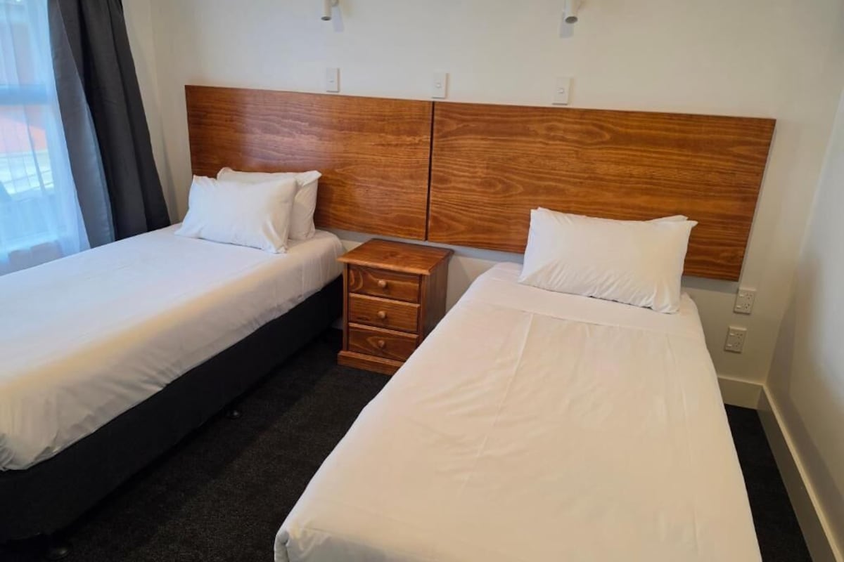A hotel room with two single beds, a wooden headboard, a small wooden nightstand, and a window with curtains.