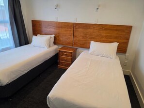 Habitación de hotel con dos camas individuales, cabecero de madera, una mesita de noche pequeña y una ventana con cortinas.