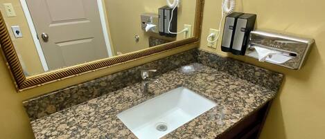 Un baño con lavamanos de encimera de granito, un espejo, un secador de pelo y un perchero para toallas.