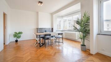 A spacious room with a wooden herringbone floor, a dining table with chairs, a potted plant, and a large window with a view of buildings.