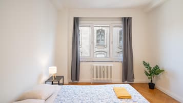 Un dormitorio con cama, mesita de noche con lámpara, radiador y ventana con cortinas.
