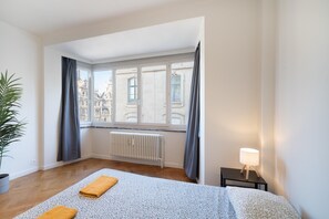 Ett hotellrum med ett stort fönster, ett element, en säng med ett blått och vitt mönstrat överkast, ett litet sängbord med en lampa och en krukväxt.
