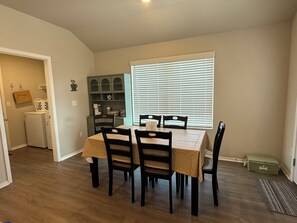 A dining area with a table and chairs, a window with blinds, and a laundry room in the background. - Ride to Bella Vista 3/4 Mi · Bike Garage · Fenced Yard, Dogs · Fireplace/grill (Pea Ridge)