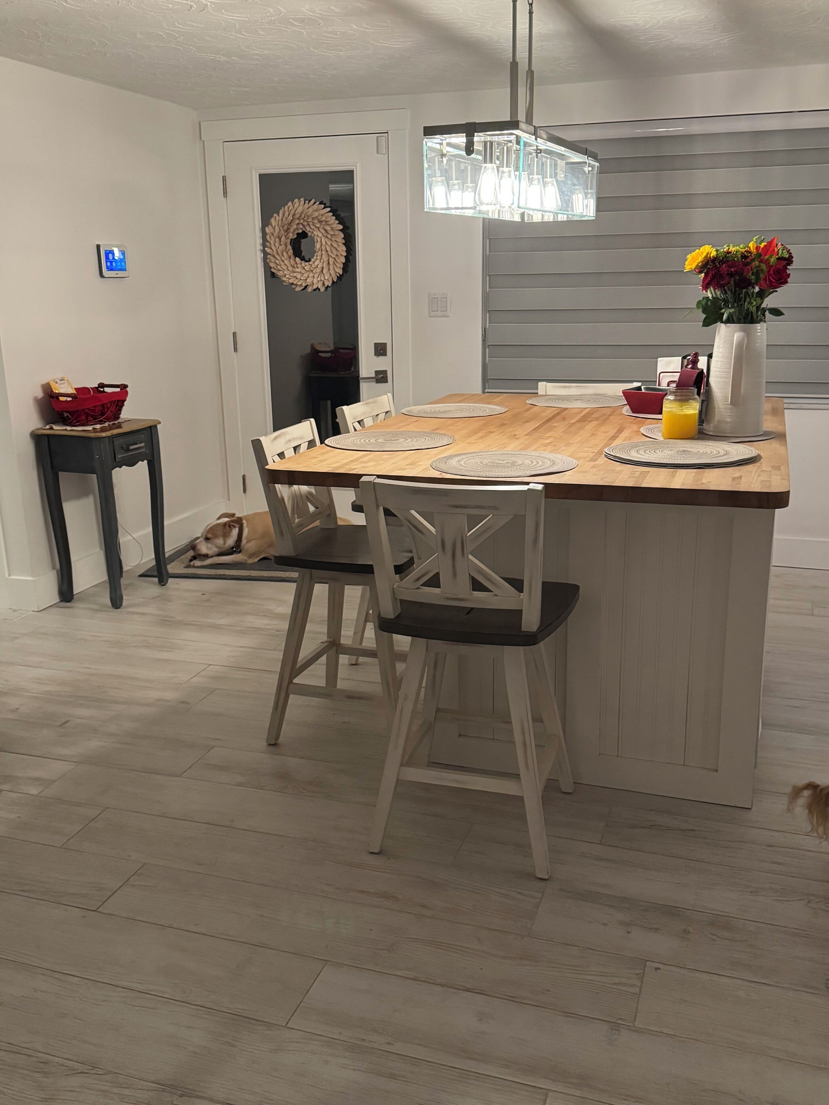 Un coin repas comprenant une table en bois, des chaises, une petite table d’appoint, une couronne décorative, une suspension et un vase avec des fleurs.