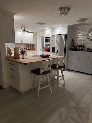 A modern kitchen with white cabinetry, a central island with bar stools, and a stainless steel refrigerator. - Beautiful beach house getaway (New Port Richey)