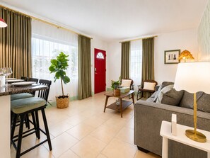 A modern living room with a sofa, a dining table set with chairs, a red door, and a large window with curtains. - Vrbo Property (Treasure Island)