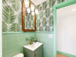 A bathroom with a green tiled wall, a white sink with a gold faucet, and a mirror with three lights. - Treasure Island Getaway | Walk to the beach | Pet-friendly (Treasure Island)