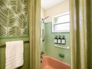 Un baño con pared de azulejos verdes, una toalla blanca, cortina de ducha, ventana y tres dispensadores de jabón.