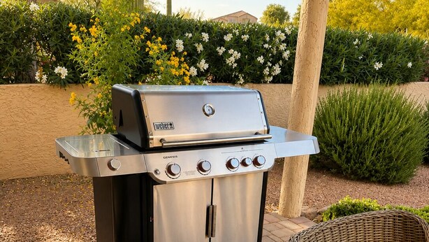 An outdoor grill with four burners, a side shelf, and a control panel, set on a brick patio with a wicker chair and table nearby.