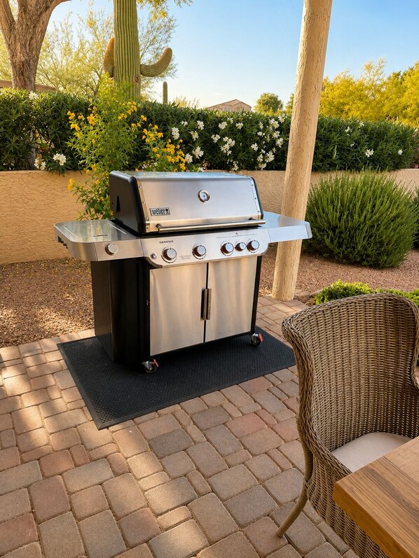 An outdoor grill with four burners, a side shelf, and a control panel, set on a brick patio with a wicker chair and table nearby.
