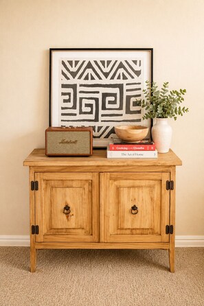 A wooden cabinet with two doors, a framed abstract artwork, a vintage radio, a bowl, a potted plant, and a book titled 'Cooking con Claudia'.