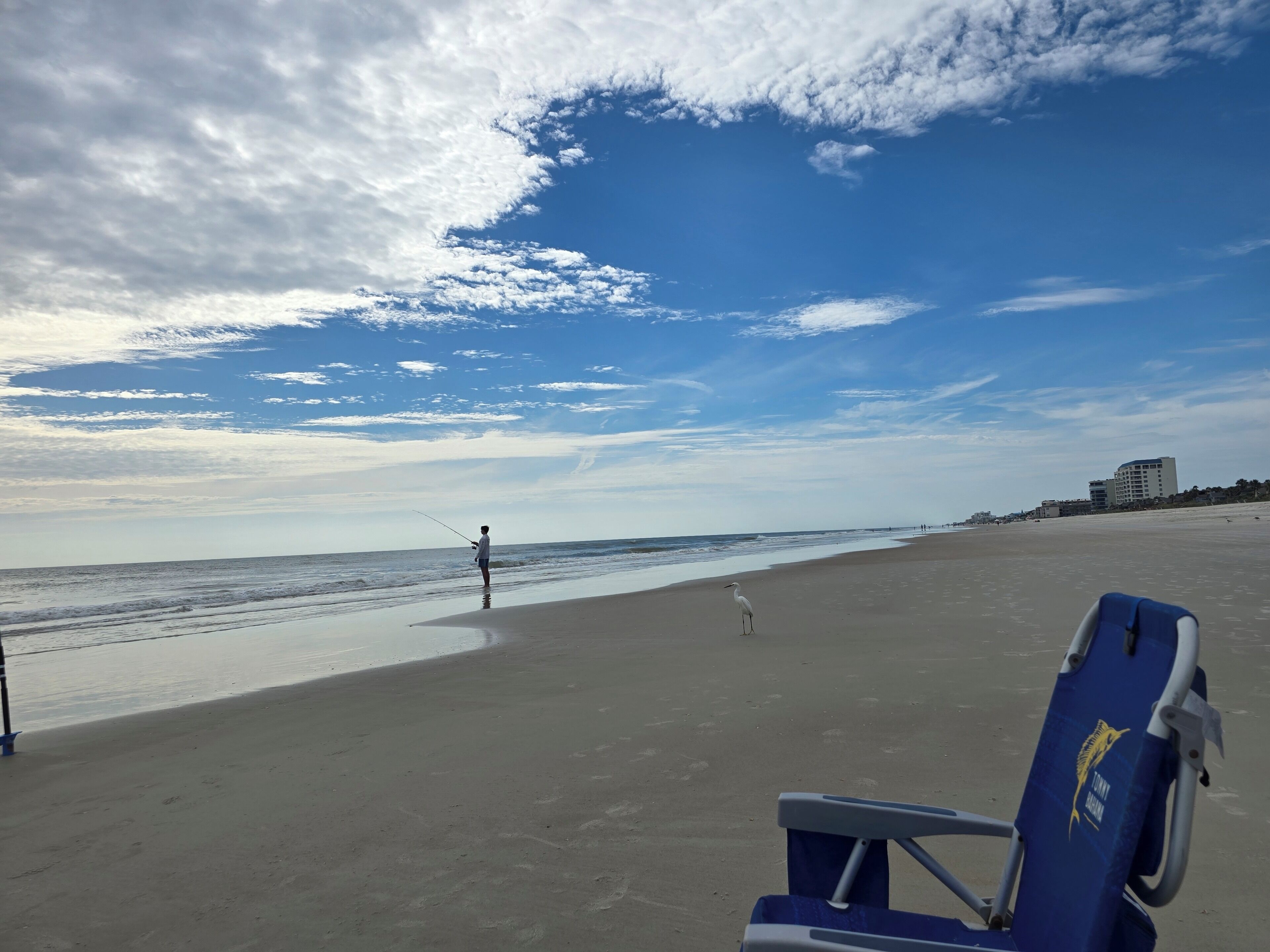 Ein Strand mit einem Stuhl und einer Angel, ein Gebäude im Hintergrund und ein klarer Himmel.