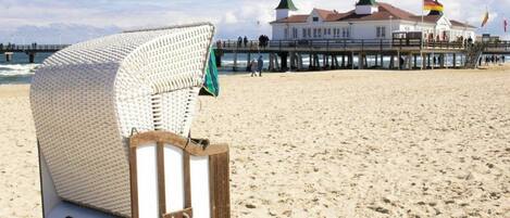 Una silla de playa con el número '80 35' sobre la arena, un muelle con edificios al fondo y banderas.