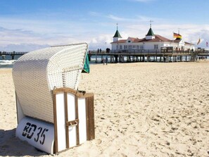 A beach chair with a number '80 35' on the sand, a pier with buildings in the background, and flags. - Holiday Apartment With 1 Bedroom (Heringsdorf)