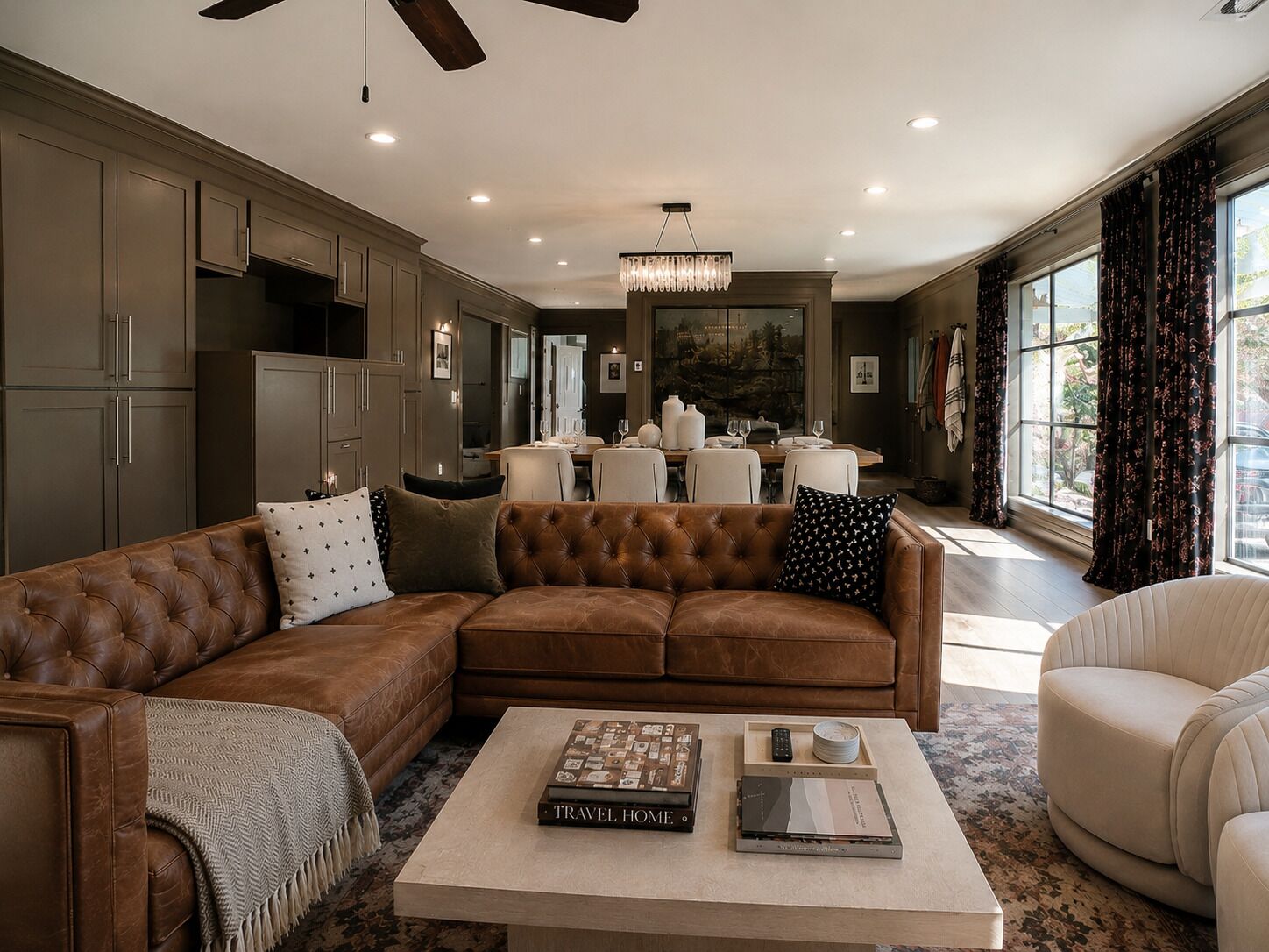 A spacious living room with a brown leather sectional sofa, a coffee table with books, and a dining area in the background.
