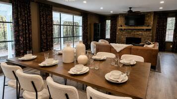A dining area with a long table set for six, surrounded by chairs, with a fireplace and a television in the background.