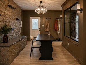 A room with a stone wall, a dark wooden table, a white chair, a chandelier, and a large window with curtains.