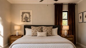 Cama bem arrumada com cobertura acolchoada, com duas mesas de cabeceira com luminárias, uma janela grande com cortinas e uma obra de arte emoldurada sobre a cama.