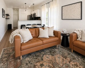 Una sala de estar moderna con un sofá de cuero marrón, un alfombra estampado y un comedor con una mesa y sillas blancas.
