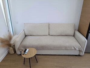 Un divano beige con due cuscini, un piccolo tavolino di legno con un telecomando e un vaso con piante essiccate.