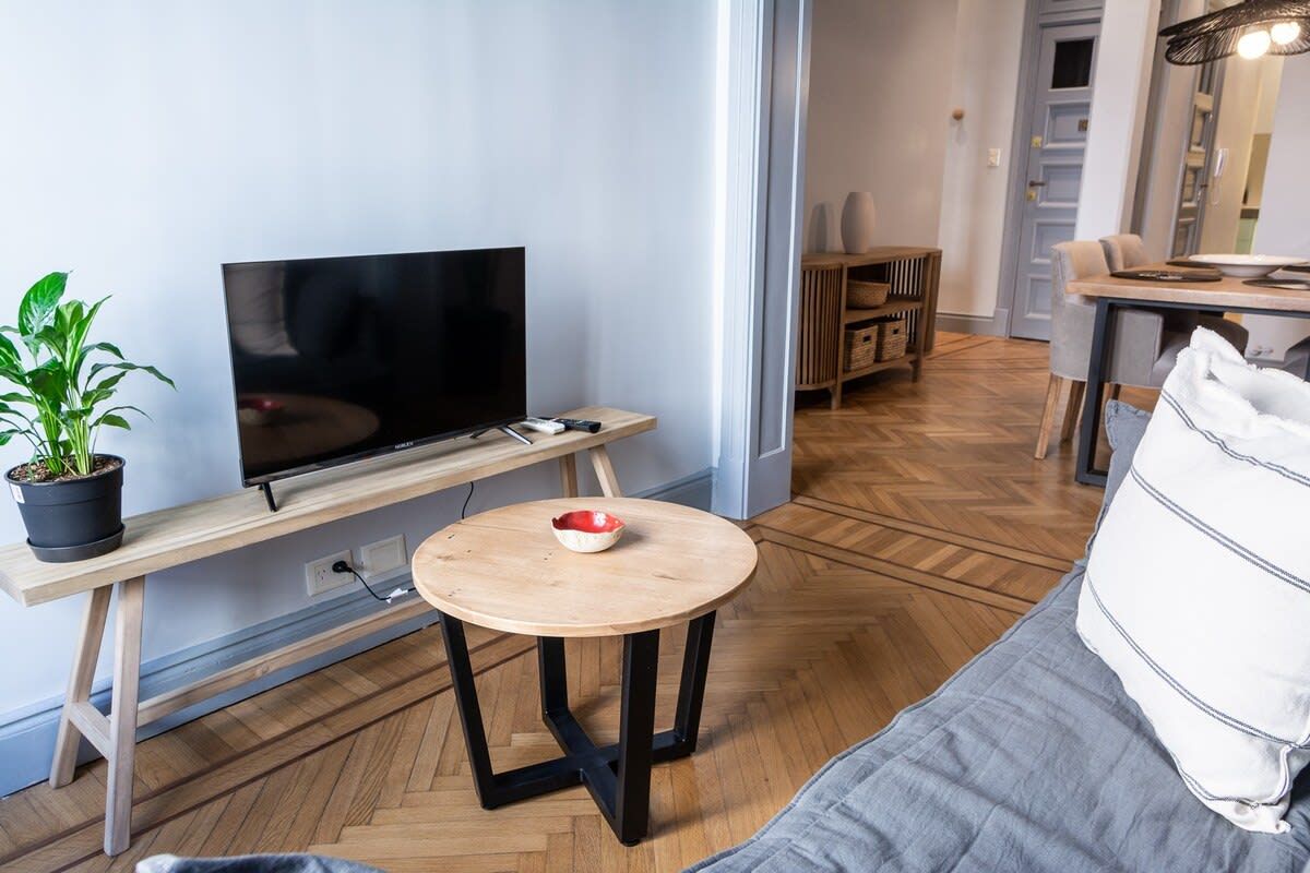 A modern living room with a flat-screen TV on a wooden stand, a round table with a bowl, a grey sofa, and a potted plant.
