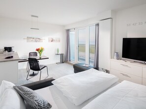 A hotel room with a bed, a desk with a chair, a television, and a balcony with a view. - Holiday Apartment in Norderney With Sauna (Norderney)