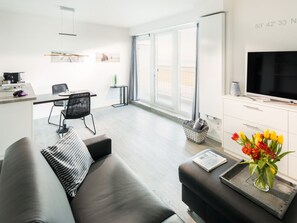 A modern living room with a sofa, a coffee table with a newspaper, a TV, and a window with a view. - Holiday Apartment in Norderney With Sauna (Norderney)