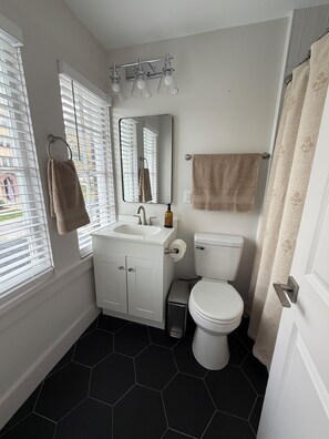 A bathroom with a white sink, mirror, and toilet, a towel rack, and a window with blinds. - 3-bedroom duplex in Dundee Omaha (Omaha)