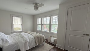 Ein Schlafzimmer mit einem Bett, einem Deckenventilator, einem Fenster mit Jalousien, einer Tür und einem kleinen Korb am Fenster.