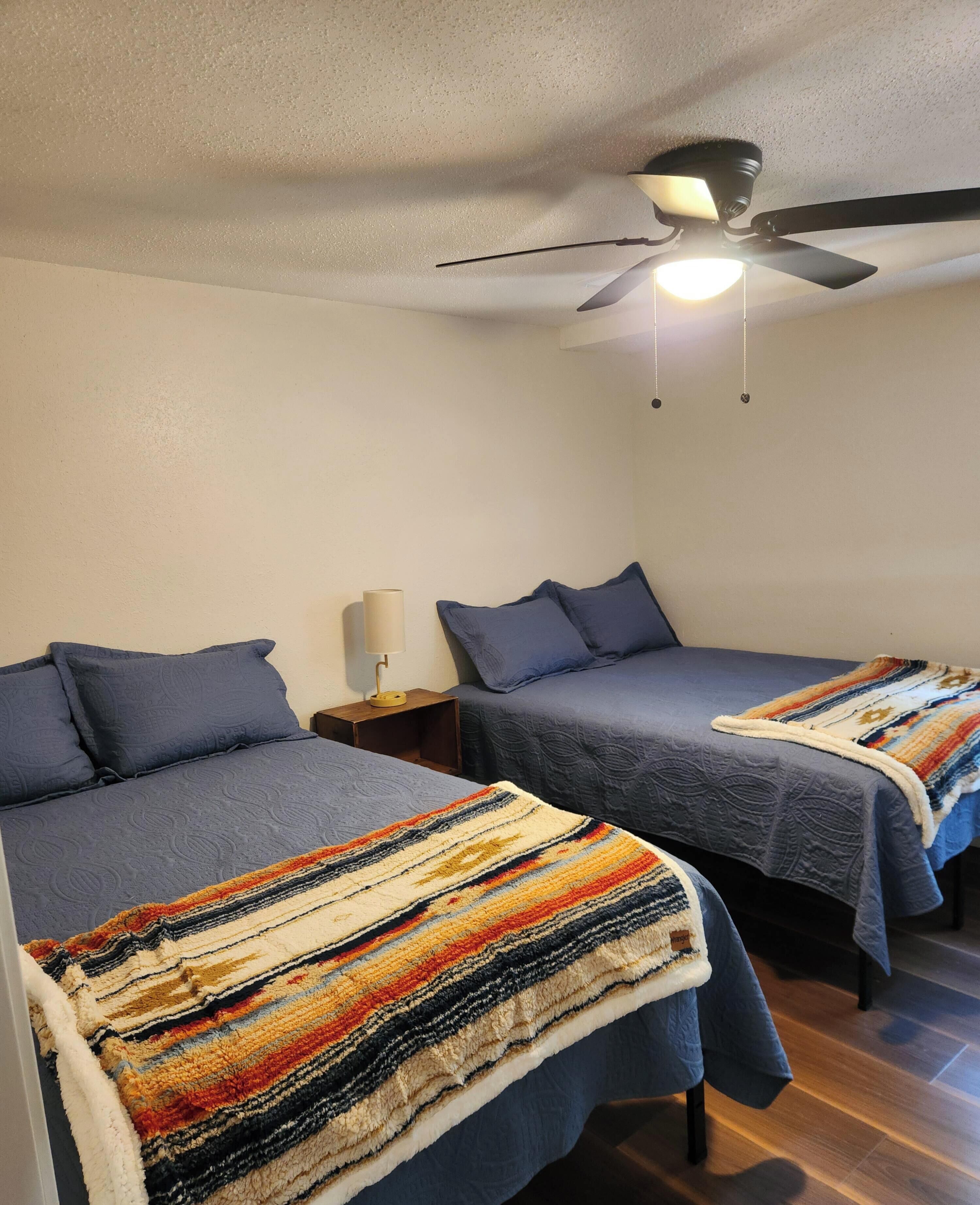 Une chambre avec deux lits, chacun recouvert d’une courtepointe bleue et agrémenté d’oreillers, une table de chevet avec une lampe et un ventilateur de plafond.