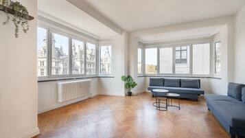 A spacious living room with large windows, a radiator, a grey sofa, and a small table.
