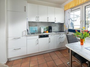 A kitchen with white cabinets, a tiled floor, and a window with yellow curtains. - Holiday Apartment in Norderney With Wi-fi (Norderney)